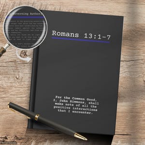 May include: A black notebook featuring the white text Romans 13:1-7 with a blue horizontal line, and a magnifying glass showing the text. Below, white text reads: For the Common Good, I, John Simmons, shall make note of all the positive interactions that I encounter. A black pen is placed diagonally across the front cover.