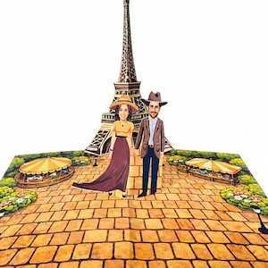 A personalized page from Our Adventure Book, a 3D pop-up book featuring a hand-illustrated Eiffel Tower. Two custom illustrated figures, created from the customer's own photos, stand together on a cobblestone path surrounded by green gardens and striped market stalls.