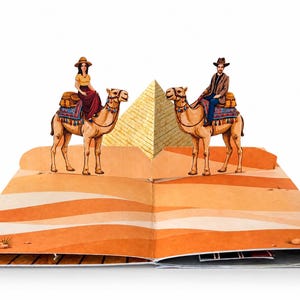 May include: A pop-up book spread featuring a three-dimensional desert scene with two camels, each carrying a rider. A large, central golden pyramid stands between the camels, set against a flat, illustrated orange and tan sand dune background.