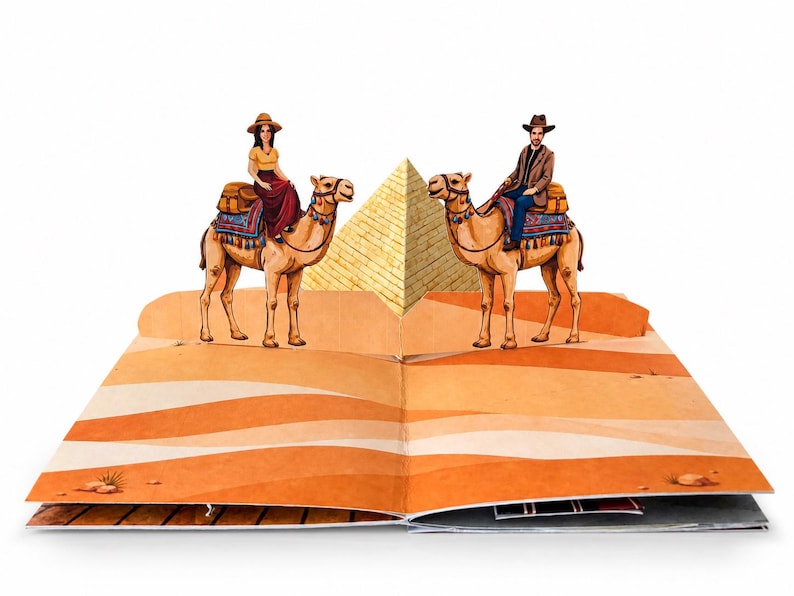 May include: A pop-up book spread featuring a three-dimensional desert scene with two camels, each carrying a rider. A large, central golden pyramid stands between the camels, set against a flat, illustrated orange and tan sand dune background.