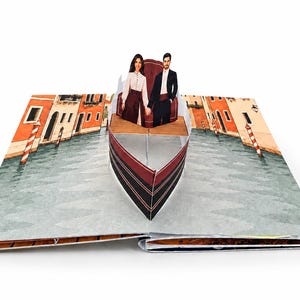 May include: A pop-up book featuring a three-dimensional paper gondola on a canal. Inside the boat, two figures stand holding hands. The background displays illustrated buildings along a waterway with red and white striped mooring poles.
