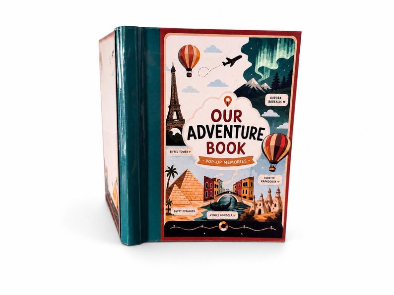 May include: A hardcover book titled Our Adventure Book Pop-Up Memories with a dark teal spine. The cover features illustrations of the Eiffel Tower, hot air balloons, the Aurora Borealis, Egyptian pyramids, and a Venice gondola.