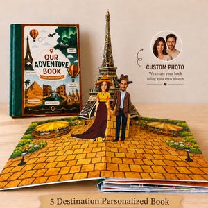 Our Adventure Book, a personalized 3D pop-up travel book with 5 destinations. The closed book cover stands beside an open page featuring a hand-illustrated Eiffel Tower, with two custom illustrated figures created from the customer's own photos standing on a cobblestone path.