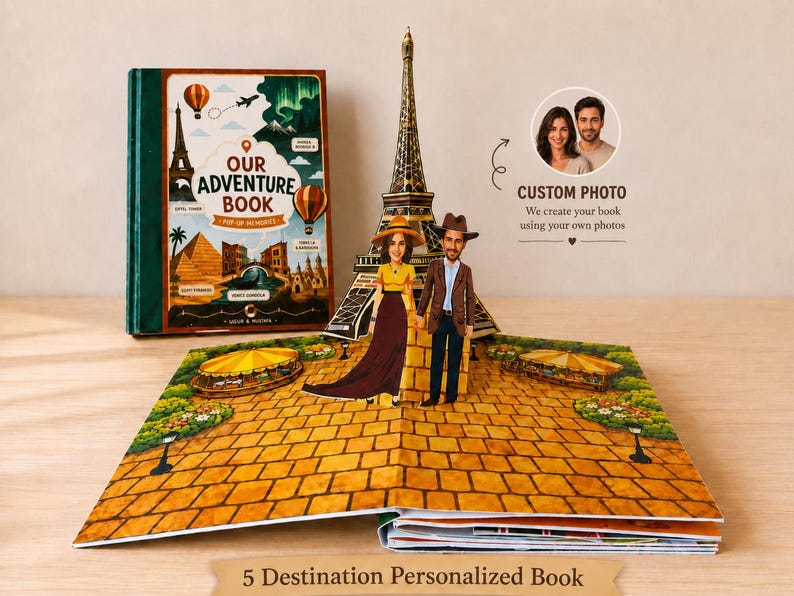 Our Adventure Book, a personalized 3D pop-up travel book with 5 destinations. The closed book cover stands beside an open page featuring a hand-illustrated Eiffel Tower, with two custom illustrated figures created from the customer's own photos standing on a cobblestone path.