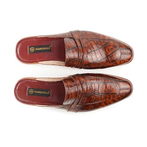 May include: A pair of brown slip-on mules featuring a textured crocodile-embossed pattern on the upper. The interior footbed is dark red with a black rectangular label displaying the gold text HANDSOLE. The shoes have a pointed toe design.