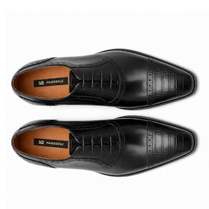 May include: A pair of black leather oxford shoes featuring a textured crocodile-embossed toe cap, brogue detailing, and black laces. The interior insole is tan and displays the black and gold text label reading HANDSOLE.