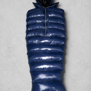 May include: A navy blue, high-gloss, quilted sleeping bag featuring a mummy-style silhouette with a hood. The exterior has a shiny, reflective finish with horizontal baffles and a central zip closure that extends from the chest to the hood.
