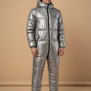 Op de afbeelding: Een metallic zilveren, eendelige gewatteerde jumpsuit met een capuchon, lange mouwen en een centrale zwarte ritssluiting. Het kledingstuk heeft een doorgestikte constructie met horizontale banen en elastische boorden bij de polsen en enkels.