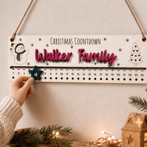 Pu&ograve; includere: Un cartello rettangolare in legno per il conto alla rovescia di Natale con il testo "Christmas Countdown" e "Walker Family" in rosa. Presenta un pupazzo di neve, un albero di Natale e un segnaposto a forma di stella verde scuro che scorre sui numeri da uno a ventiquattro.