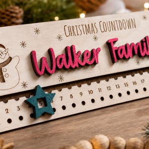 Pu&ograve; includere: Un cartello rettangolare in legno per il conto alla rovescia di Natale con il testo inciso "CHRISTMAS COUNTDOWN" e un pupazzo di neve. Il cartello presenta la scritta rosa in rilievo "Walker Family" e una stella mobile color ottanio.