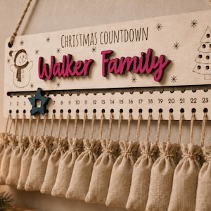 Pu&ograve; includere: Un cartello rettangolare in legno per il conto alla rovescia di Natale con il testo "CHRISTMAS COUNTDOWN" e "Walker Family" in corsivo rosa. Include un pupazzo di neve, un albero di Natale, una traccia numerata da 1 a 24 con un segnaposto a stella blu e ventiquattro piccoli sacchetti di tessuto beige appesi a spaghi di juta.