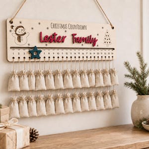Pu&ograve; includere: Un cartello rettangolare in legno per il conto alla rovescia di Natale con la scritta "Christmas Countdown" e "Lester Family" in rosso. Presenta un pupazzo di neve, un albero, i numeri da 1 a 24, una stella blu e due file di sacchetti in tessuto beige.