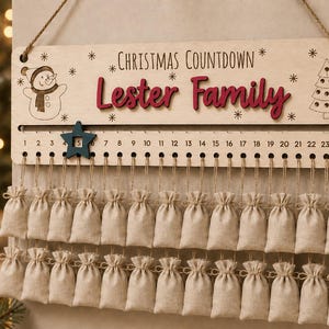 Pu&ograve; includere: Un cartello in legno per il conto alla rovescia di Natale con la scritta "Christmas Countdown" e "Lester Family" in rosso. Presenta un pupazzo di neve, un albero di Natale e i numeri da 1 a 24 con un segnaposto a stella blu. Ventiquattro sacchetti in tessuto beige pendono sotto.