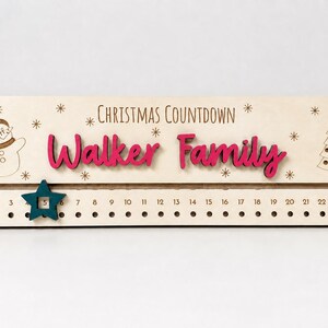 Pu&ograve; includere: Un cartello rettangolare in legno chiaro per il conto alla rovescia di Natale con il testo inciso "CHRISTMAS COUNTDOWN" e la scritta in rilievo rosa scuro "Walker Family". Include un pupazzo di neve, un albero di Natale e un segnaposto a stella color ottanio.