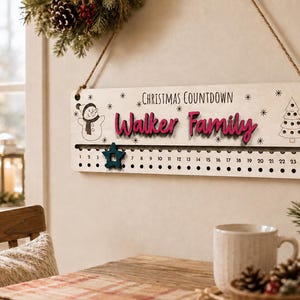 Pu&ograve; includere: Un cartello rettangolare in legno per il conto alla rovescia di Natale con una corda di iuta. Il cartello presenta la scritta "Christmas Countdown" e "Walker Family" in rosso, con un pupazzo di neve, un albero di Natale e i numeri da 1 a 24 con una stella blu.