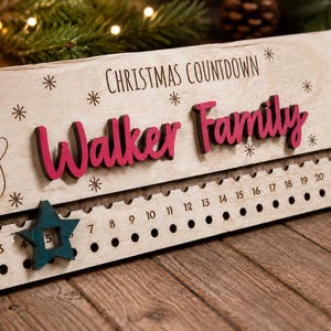 Pu&ograve; includere: Un cartello rettangolare in legno per il conto alla rovescia di Natale con il testo "CHRISTMAS COUNTDOWN" e "Walker Family" in lettere rosa in rilievo. Include un pupazzo di neve inciso al laser, un albero di Natale, fiocchi di neve e una traccia numerata da 1 a 24 con una stella mobile color verde acqua.