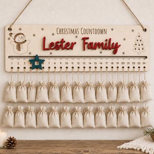 Pu&ograve; includere: Un cartello in legno per il conto alla rovescia di Natale con la scritta Lester Family in rosso, un'incisione di un pupazzo di neve e un albero, e un segnaposto a forma di stella blu. Ventiquattro sacchetti in tessuto beige pendono da una corda.