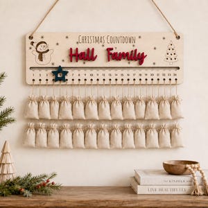 Pu&ograve; includere: Un cartello per il conto alla rovescia di Natale in legno chiaro con il testo "Christmas Countdown" e "Hall Family" in rosso. Presenta un'illustrazione di un pupazzo di neve e un albero, una traccia numerata da 1 a 24 con un segnaposto a stella blu scuro e due file di piccoli sacchetti di tessuto beige.
