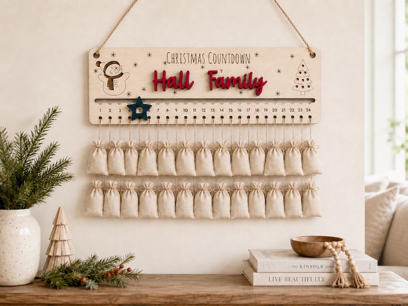Pu&ograve; includere: Un cartello per il conto alla rovescia di Natale in legno chiaro con il testo "Christmas Countdown" e "Hall Family" in rosso. Presenta un'illustrazione di un pupazzo di neve e un albero, una traccia numerata da 1 a 24 con un segnaposto a stella blu scuro e due file di piccoli sacchetti di tessuto beige.