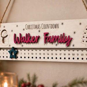 Pu&ograve; includere: Un cartello rettangolare in legno per il conto alla rovescia di Natale con il testo "CHRISTMAS COUNTDOWN" e "Walker Family" in corsivo rosa. Include un pupazzo di neve, un albero di Natale e una stella scorrevole color ottanio.