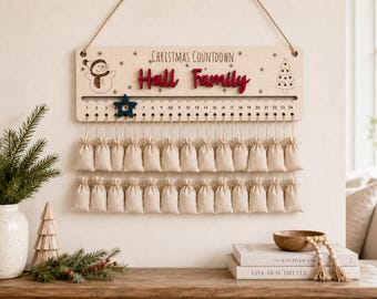 Può includere: Un cartello per il conto alla rovescia di Natale in legno chiaro con il testo "Christmas Countdown" e "Hall Family" in rosso. Presenta un'illustrazione di un pupazzo di neve e un albero, una traccia numerata da 1 a 24 con un segnaposto a stella blu scuro e due file di piccoli sacchetti di tessuto beige.
