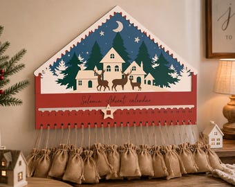 Può includere: Un calendario dell'avvento in legno a forma di casa con base rossa e una scena notturna blu con case bianche, pini, una luna e cervi. Include la scritta Solomia Advent calendar, i numeri da 1 a 24, un segnaposto a stella e ventiquattro piccoli sacchetti di iuta.