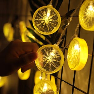May include: A string of decorative LED lights featuring translucent yellow circular covers shaped like lemon slices. Each light emits a warm glow, connected by a clear wire. The circular lemon slice covers have a textured, radial pattern.