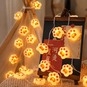 Puede incluir: Una cadena de luces LED con cubiertas en forma de huellas de animales en color blanco y naranja. Cada cubierta individual tiene forma de huella con un centro brillante. Las luces están conectadas por un cable fino y emiten una luz cálida.