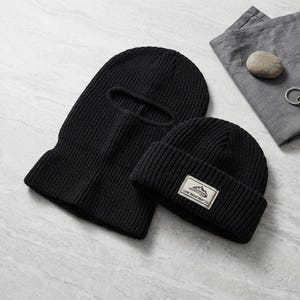 May include: A black knitted balaclava with a single eye opening and a matching black knitted beanie featuring a rectangular patch with a mountain graphic and the text "OUTDOOR GEAR" printed in black on a light-colored background.