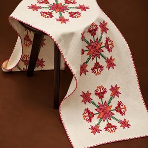 Puede incluir: Un camino de mesa rectangular de color blanco roto con bordados geométricos repetidos en forma de estrella en hilo rojo, naranja y verde. La tela tiene un borde cosido en rojo y está drapeada sobre un taburete de madera oscura.