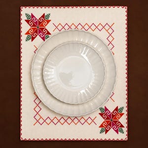 Puede incluir: Un mantel individual rectangular de color crema con un borde geométrico en punto de cruz rojo y naranja y motivos de estrellas en las esquinas en rojo, rosa y verde. Dos platos de cerámica blanca con bordes festoneados.
