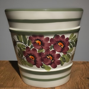 May include: A white ceramic flower pot featuring horizontal sage green stripes around the top and bottom. The center is decorated with a hand-painted floral motif consisting of dark purple blossoms with orange centers and green leaves.