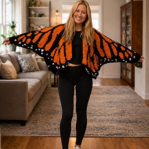 May include: A crochet cardigan featuring a vibrant monarch butterfly wing pattern in bright orange, black, and white. The garment has wide, open sleeves and a draped silhouette, worn over a black crop top, black leggings, and white sneakers.