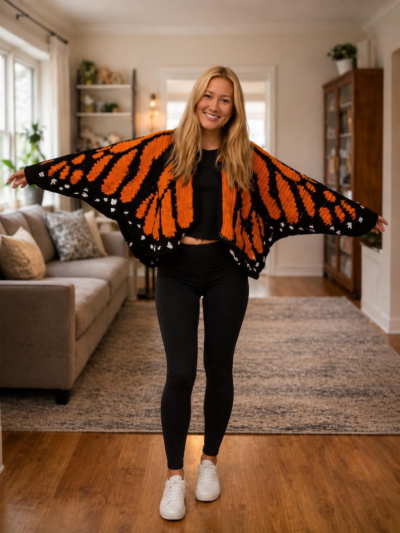 May include: A crochet cardigan featuring a vibrant monarch butterfly wing pattern in bright orange, black, and white. The garment has wide, open sleeves and a draped silhouette, worn over a black crop top, black leggings, and white sneakers.