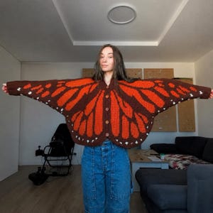 May include: A long-sleeve knit cardigan featuring a monarch butterfly wing pattern in dark brown and vibrant orange, with small white accents along the edges. The garment has a button-down front with four brown buttons and a wide, wing-like silhouette.