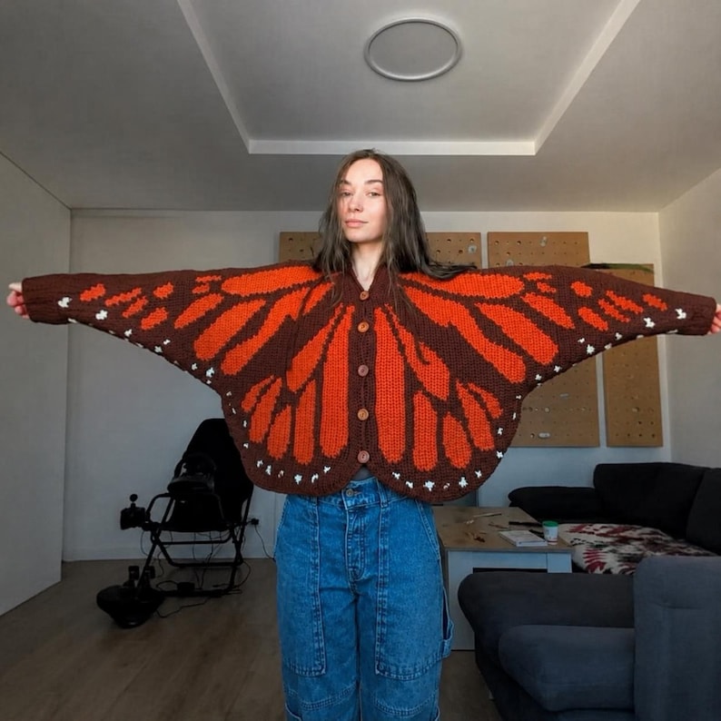 May include: A long-sleeve knit cardigan featuring a monarch butterfly wing pattern in dark brown and vibrant orange, with small white accents along the edges. The garment has a button-down front with four brown buttons and a wide, wing-like silhouette.