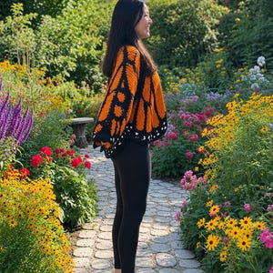 May include: A crochet sweater featuring a vibrant orange and black monarch butterfly wing pattern with white accents along the hem, paired with solid black leggings and white sneakers, worn in a profile view against a garden setting.