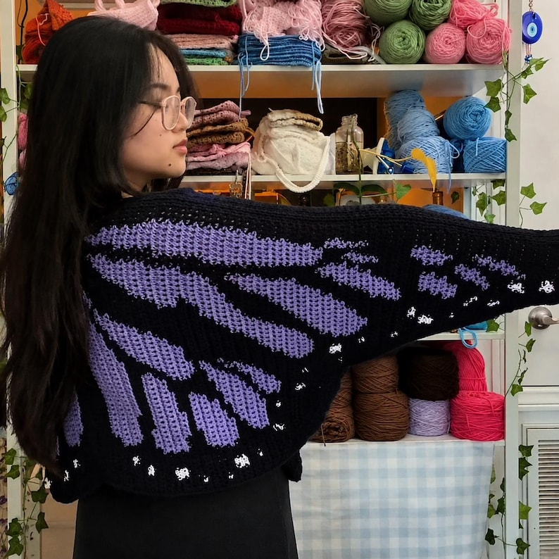 May include: A black and purple crocheted cardigan featuring a butterfly wing pattern. The garment has a dark base with vibrant purple sections and small white accents along the bottom edge, designed with wide sleeves and a cropped silhouette.