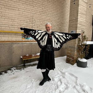 May include: A black and white butterfly wing patterned cardigan with red accents, worn over a black shirt and a black kilt. The cardigan features wide, wing-shaped sleeves that extend outward, creating a distinct silhouette against the snowy background.