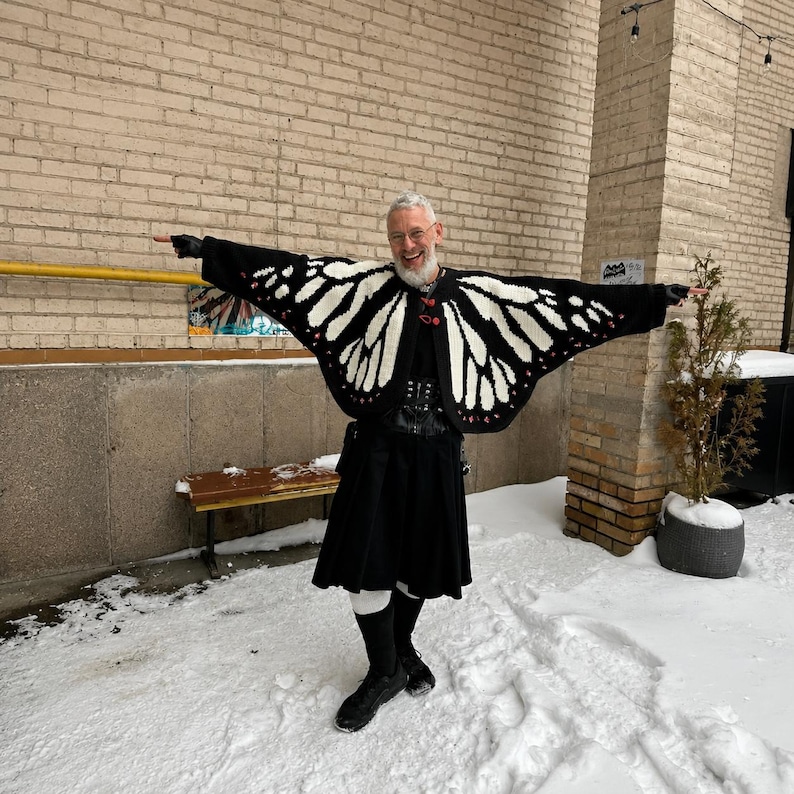 May include: A black and white butterfly wing patterned cardigan with red accents, worn over a black shirt and a black kilt. The cardigan features wide, wing-shaped sleeves that extend outward, creating a distinct silhouette against the snowy background.