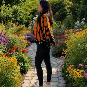 May include: A crochet cardigan featuring a vibrant orange and black monarch butterfly wing pattern with a black border and small white accents, paired with solid black leggings and white sneakers, worn in a garden setting.