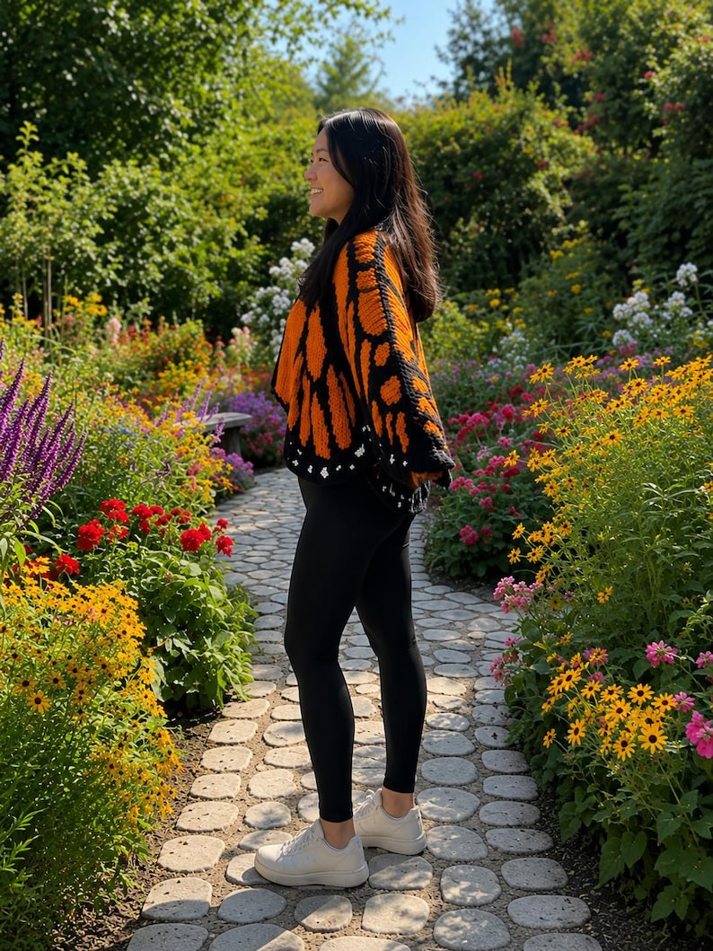 May include: A crochet cardigan featuring a vibrant orange and black monarch butterfly wing pattern with a black border and small white accents, paired with solid black leggings and white sneakers, worn in a garden setting.