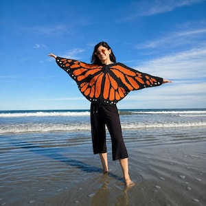 May include: A black and orange crochet cardigan featuring a monarch butterfly wing pattern with a zip-up front, worn with black cropped trousers and round-tinted sunglasses. The garment has wide, wing-like sleeves that extend when arms are outstretched.
