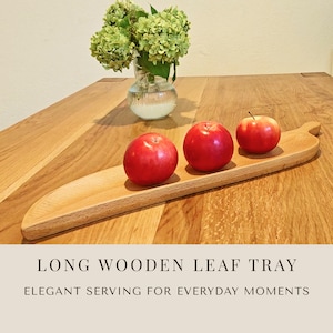 Op de afbeelding: Een lang, lichtgekleurd houten dienblad in de vorm van een blad, met drie felrode appels. Het dienblad heeft een gladde afwerking met natuurlijke houtnerf en een taps toelopend uiteinde. De tekst "LONG WOODEN LEAF TRAY" en "ELEGANT SERVING FOR EVERYDAY MOMENTS" is zichtbaar.