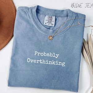 May include: A blue jean coloured short-sleeve t-shirt with a crew neckline. The shirt features the white text 'Probably Overthinking' printed on the front. A small gold-toned pendant necklace rests on the collar area of the garment.