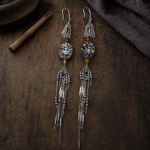May include: A pair of long dangle earrings featuring silver-tone hooks, small silver chains, and amber-colored beads. Each earring includes a central oval-shaped white bead with a black crackle pattern, finished with multiple hanging silver chains.