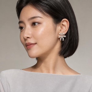 May include: A silver-toned stud earring featuring a double bow design. The upper bow is smaller and sits above a larger lower bow, with a small clear stone set at the center of each bow. The earring is worn on the earlobe.