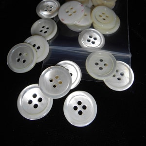 May include: A collection of white, round buttons with four holes, some loose and some in a clear plastic bag. The buttons have a pearlescent sheen and vary slightly in size. The background is black.