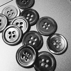 May include: A collection of dark brown buttons with four holes each, scattered on a gray surface. The buttons vary slightly in shade and size, creating a textured appearance. The image is in black and white.