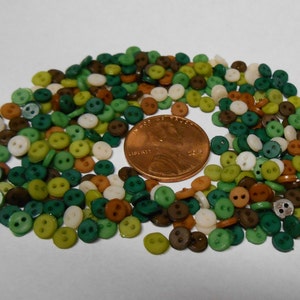 Puede incluir: Un gran surtido de botones pequeños de dos agujeros en colores verde, marrón y blanco. Los botones están esparcidos sobre una superficie blanca, con una moneda de un centavo de dólar estadounidense para la escala.